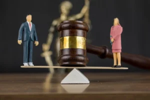 Two figurines depicting a couple stand on opposite ends of a balance scale with a judge’s gavel between them, representing the legal process of divorce proceedings in Singapore